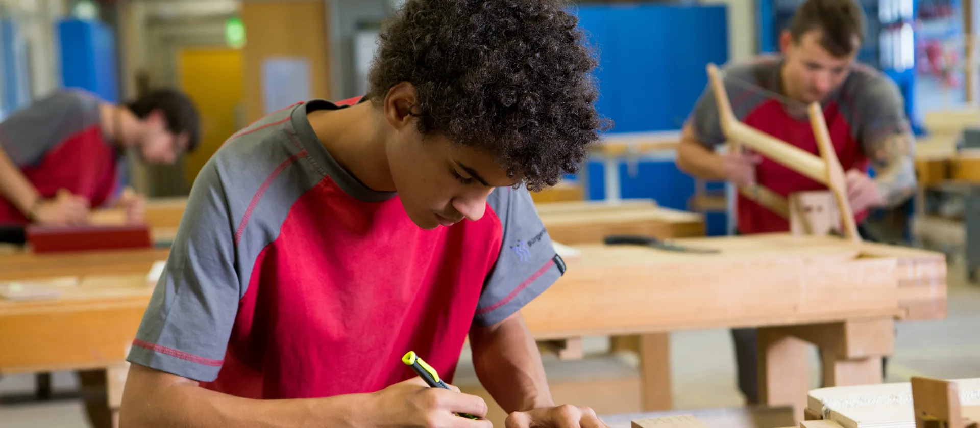 Eine Person, die gerade an einer Wrkbank arbeitet. Im Hintergrund sieht man unscharf zwei weitere Mitarbeitende.